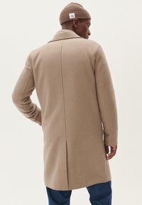 Beige wollen jas met een getailleerde pasvorm, achternaaddetail en lange mouwen. Gecombineerd met een bruine geribde gebreide beanie en blauwe denim broek.