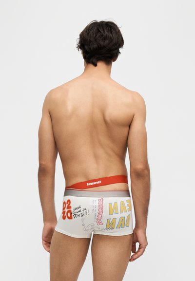 Νέος άνδρας με γυμνή πλάτη που φοράει λευκά boxer briefs με πολύχρωμο κείμενο και κόκκινη ζώνη, στέκεται μπροστά σε απλό φωτεινό φόντο.