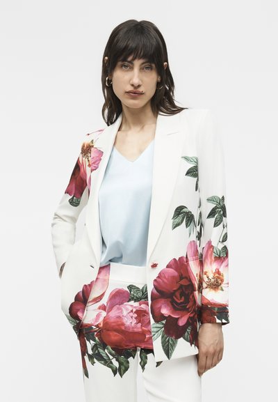 Γυναίκα που φοράει λευκό κοστούμι με μεγάλο κόκκινο και ροζ floral μοτίβο, ανοιχτό γαλάζιο πουκάμισο από κάτω, στέκεται με το ένα χέρι στην τσέπη.