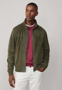 Veste bomber en suède vert olive avec col officier, fermeture éclair à l'avant et poches latérales, associée à un pull bordeaux et un pantalon blanc.
