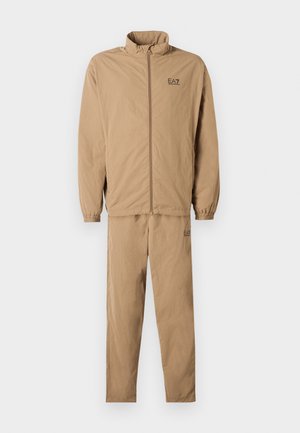 Beige Jogginganzug-Set mit einer Reißverschlussjacke mit hohem Kragen und elastischen Bündchen, kombiniert mit passenden Hosen, beide mit dem EA7-Logo.