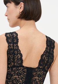 Mustakankainen bodysuit, jossa on kukkakuvioita, leveät pitsihihnat ja matala selkäosa, joka esittelee yksityiskohtaista tekstuuria ja monimutkaista ompelua.