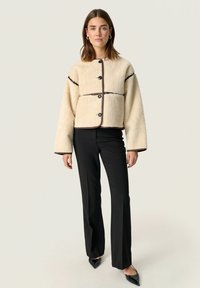 Giacca in finta pelliccia shearling crema con design corto, bordo marrone a contrasto, grandi bottoni neri e maniche ampie. Abbinata a pantaloni neri.