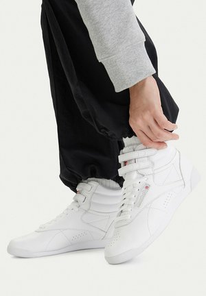 Person, die die Manschette schwarzer Hosen anpasst, trägt weiße High-Top-Sneaker mit doppelten Riemen, wobei ein Ärmel eines grauen Sweatshirts sichtbar ist.