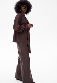 Kobieta z kręconymi włosami i okularami, ubrana w luźną brązową bluzkę z bufiastymi rękawami oraz szerokie brązowe spodnie, stoi bokiem.