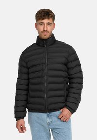 Sort pufferjakke med høj krave, vandret quiltet design, lynlås foran og ribbede manchetter. Båret over lyseblå jeans.