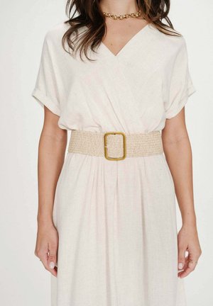 Femme portant une robe portefeuille blanche à manches courtes, accompagnée d'une large ceinture tissée beige avec une grande boucle carrée dorée.