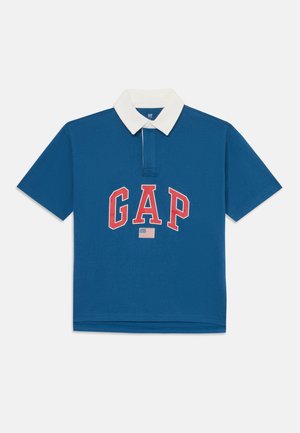 Cămașă polo albastră cu guler alb, cu "GAP" scris în litere roșii texturate și un mic detaliu cu steagul SUA dedesubt. Mâneci scurte, din amestec de bumbac.