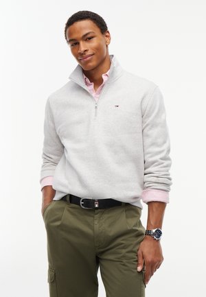 Junger Mann trägt einen hellgrauen Quarter-Zip-Pullover über einem rosa Hemd und olivgrünen Hosen mit schwarzem Gürtel und Armbanduhr.