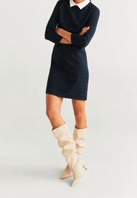 Abito a quadri blu navy con colletto bianco, maniche lunghe e silhouette aderente, abbinato a stivali a metà polpaccio, crema, morbidi e con tacco.
