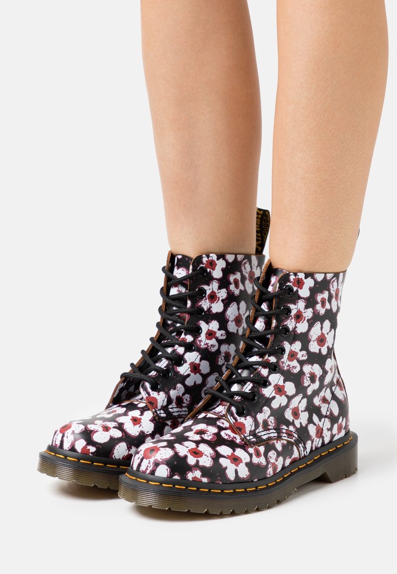 Jambes portant des bottes noires à lacets avec un motif floral blanc et rouge et une couture jaune sur des semelles épaisses translucides sur fond blanc.