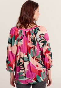 Blusa floreale con maniche lunghe a sbuffo, scollatura arricciata e motivi di foglie nei colori vivaci di rosa, rosso e verde su uno sfondo chiaro.