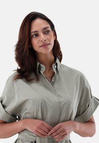 Camicia oversize verde chiaro con colletto e maniche corte arrotolate, dotata di una cintura da legare in vita. Tessuto in cotone misto liscio.