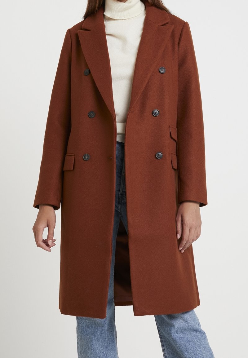 Brun uldblandet dobbeltbrystet frakke med sorte knapper, slagkrave og sidelommer, parret med en cremefarvet turtleneck og blå jeans.
