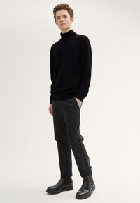 Schwarzer Strick-Turtleneck-Pullover, eng anliegende schwarze Hose und schwarze Stiefeletten mit seitlichen Gummizügen, stehend vor einem hellen Hintergrund.