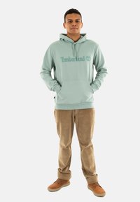 Felpa verde menta con logo 'Timberland', tasca a marsupio frontale, abbinata a pantaloni in corduroy beige e scarpe color terra, in piedi su uno sfondo bianco.