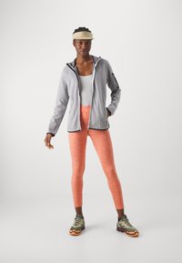 Gray zip-up jacket with black accents, white tank top, bright orange leggings with textured pattern, and green athletic shoes with orange soles.