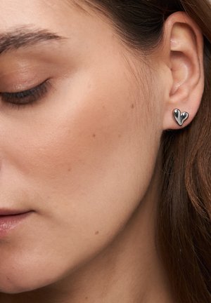 Nahaufnahme des Gesichts einer Frau, die einen silbernen herzförmigen Ohrstecker an ihrem Ohr trägt und einen ruhigen, neutralen Gesichtsausdruck zeigt.