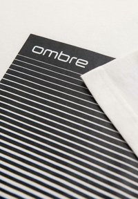 Bílý bavlněný tričko částečně složené přes černobílou pruhovanou látku s nápisem "ombre" vytištěným moderním písmem v horním levém rohu.