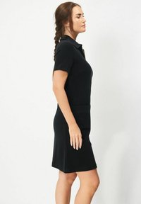 Svart polo-stil klänning med korta ärmar, en figursydd bodice och rak nederkant. Inkluderar en ficka på framsidan för extra detaljer.