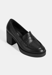 Zwarte leren loafers met blokhak en een textuur bovenwerk, voorzien van een voorband en een ronde neus. De zool is plat en van rubber.