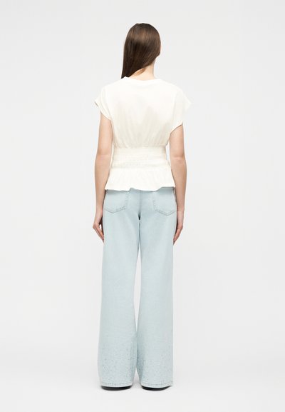 Γυναίκα που στέκεται με την πλάτη γυρισμένη, φορώντας λευκό μπλουζάκι με κοντά μανίκια και ζωνάκι στη μέση, και ανοιχτόχρωμα μπλε τζιν παντελόνια με φαρδιά μπάτζα και διακριτικές διακοσμήσεις.