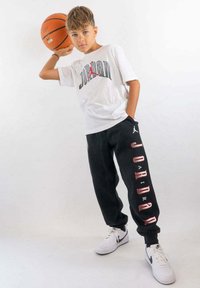 Witte katoen t-shirt met "Jordan" logo, zwarte sweatpants met "Jordan" tekst en witte Nike sneakers. Houdt een basketbal vast.