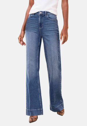 Frau trägt hoch-taillierte, weit geschnittene blaue Jeans mit umgekrempelten Säumen, kombiniert mit spitz zulaufenden Pumps mit Schlangenmuster und einem weißen ärmellosen Oberteil.