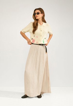 Frau mit Sonnenbrille, besticktem kurzärmligen Hemd mit Knöpfen, plissiertem langem beigen Rock, schwarzem Gürtel und schwarzen Schuhen, stehend mit den Händen in der Hüfte.