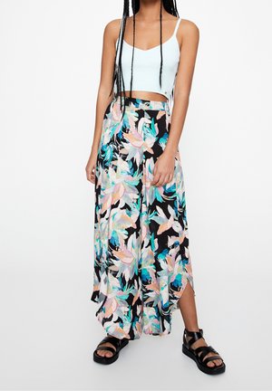 Kobieta ubrana w biały krótki top na ramiączkach, szerokie spodnie w kwiatowy wzór oraz czarne sandały na platformie, stojąca na jednolitym tle.