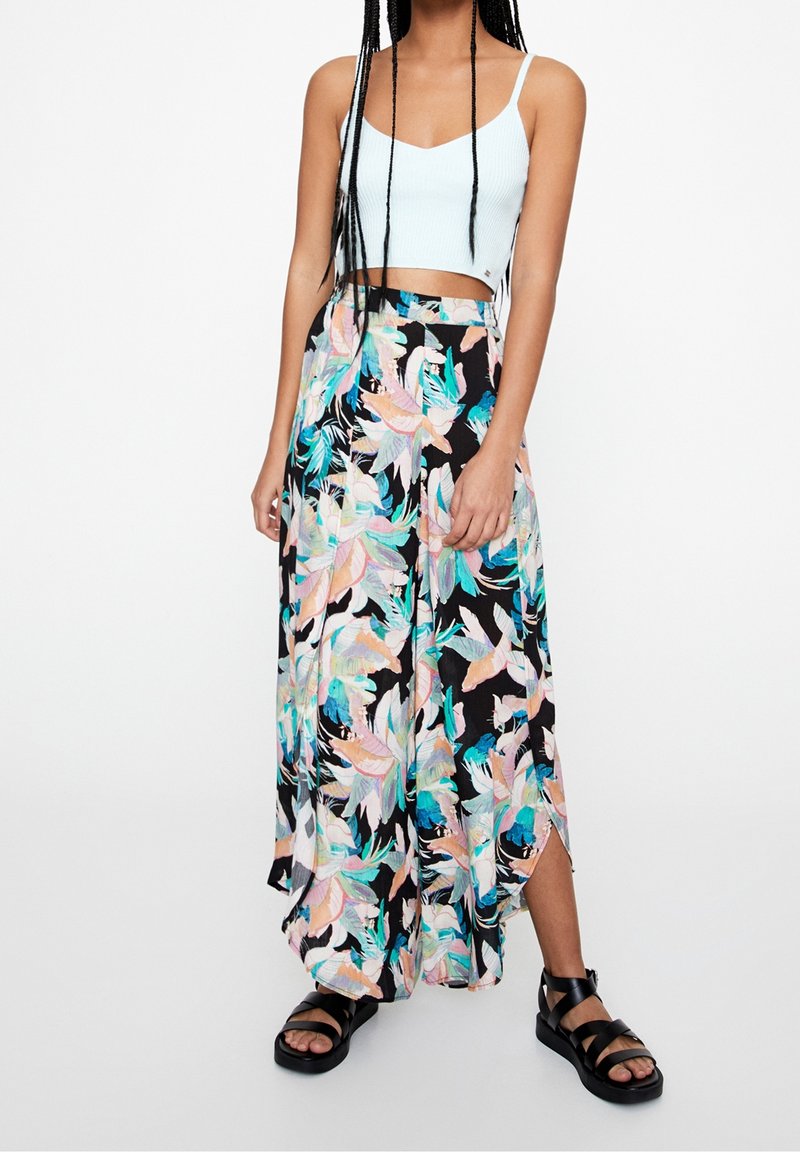 Femme portant un débardeur blanc court, un pantalon large à imprimé floral, et des sandales plateformes noires, debout devant un fond uni.