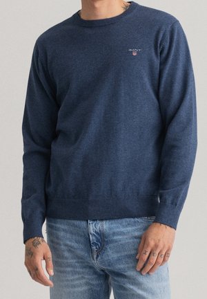 Mężczyzna w ciemnoniebieskim swetrze GANT z długim rękawem i jasnoniebieskich jeansach, pokazujący tatuaże na rękach oraz część tatuażu na szyi.