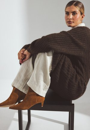 Suéter de punto acanalado marrón, pantalones de pierna ancha color crema y botas de ante marrones con punta afilada, sentado en una silla negra contra un fondo claro.
