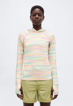 Ember pasztell csíkos, testhez simuló, kapucnis, hosszú ujjú felsőt és világoszöld rövidnadrágot viselve, egyszínű fehér háttér előtt áll.