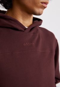 Tummanpunainen huppari pehmeällä pinnalla, jossa on kengurupussi ja brodeerattu Adidas-logo rinnan ylle. Huppu on kiinnitetty saumattomalla muotoilulla.