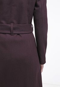 Burgundská vlněná bunda s páskem v pase, přizpůsobený střih, dlouhé rukávy a čisté linie, která prezentuje hladkou texturu a minimalistický design.