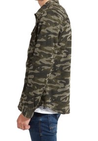 Giacca camo in tonalità verde e grigio. Realizzata in tessuto testurizzato, presenta una chiusura anteriore con bottoni e cuciture visibili. Le maniche possono essere arrotolate fino ai polsini.