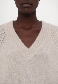 Gros plan d'une personne portant un pull en maille duveteuse beige clair à col en V, montrant le cou, la clavicule et le bas du visage.