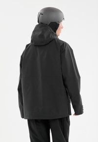 Svart vattentät jacka med huva, långa ärmar och sidodragkedjor, ihopparad med en grå hjälm, visad bakifrån mot en neutral bakgrund.