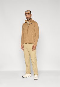 Bēša vilnas jaka ar rāvējslēdzēju un sānu kabatām; bēšas bikses ar regulējamām manšetēm. Baltas sporta kurpes un bēša cepure ar logotipu. Ikdienas stils.