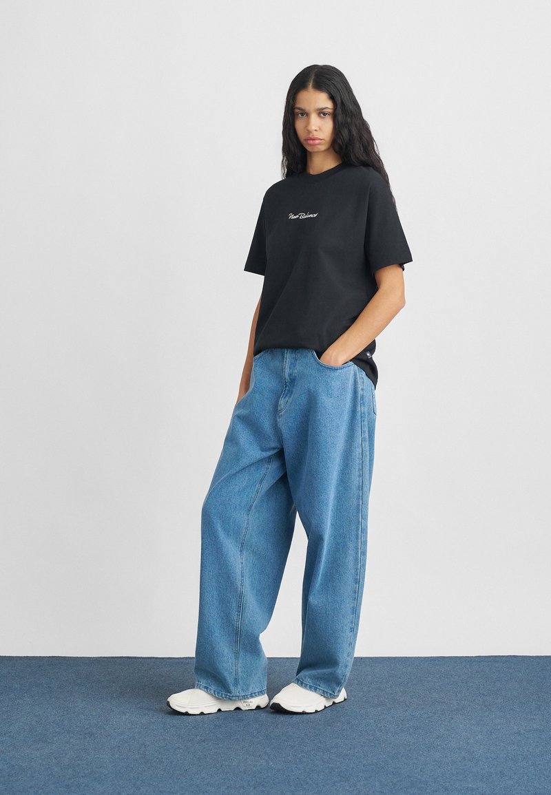 T-shirt di cotone nera con logo ricamato, abbinata a jeans in denim azzurro chiaro larghi. Il modello indossa sneakers bianche con dettagli neri.