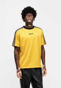 Κίτρινο μπλουζάκι adidas με μαύρες λεπτομέρειες και ρίγες στα μανίκια. Διαθέτει στρογγυλή λαιμόκοψη και ανάγλυφο μοτίβο στο ύφασμα.