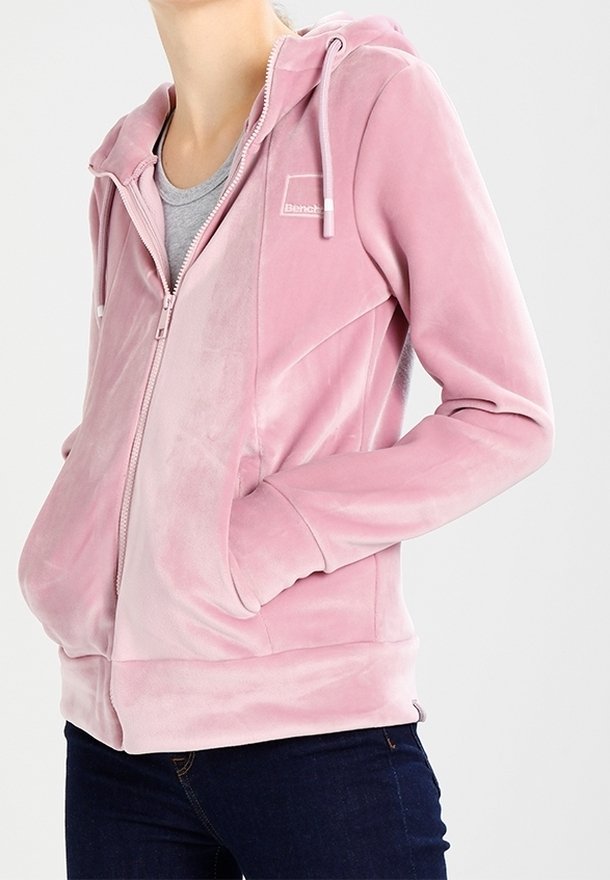 Kvinna klädd i en rosa zip-up huvtröja med huva och framfickor, i kombination med mörkblå jeans och en grå t-shirt under.