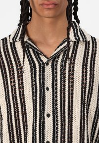 Camisa de manga corta en crema y negro, con un patrón a rayas y un tejido texturizado, cuello abierto y botones negros.