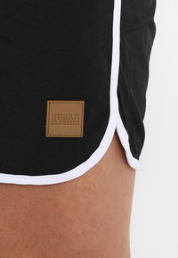 Pantaloni scurți negri cu margini albe și o etichetă rectangulară din piele maro, inscripționată cu "URBAN CLASSICS". Materialul pare să fie ușor și texturat.
