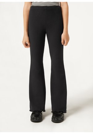 Persona con pantalones acampanados negros de canalé con dobladillo ondulado y zapatos de plataforma negros, de pie contra un fondo liso.