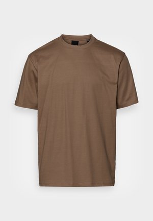 Homme portant un t-shirt à manches courtes vert olive rentré dans un jean écru avec une ceinture noire, debout avec une main dans la poche.