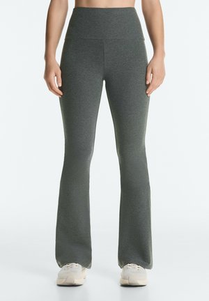 Persoon draagt donkergrijze, hooggetailleerde uitlopende legging en witte sneakers, staand tegen een effen lichte achtergrond.