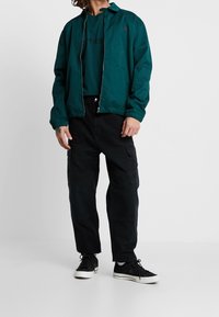 Teal bomberjack met een ritssluiting aan de voorkant, zwarte cargo broeken en zwarte sneakers. Het ontwerp heeft elastische manchetten en een eenvoudig logo.