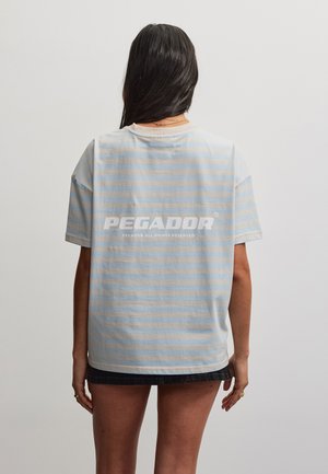 Person med langt mørkt hår står med ryggen til iført en lyseblå og hvide stribet T-shirt med "PEGADOR" i store hvide bogstaver på ryggen.